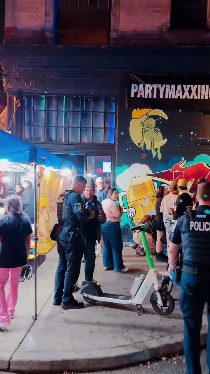 Two Seattle Police officers talking with people on a crowded Capitol Hill sidewalk at night — a Lime e-scooter in the foreground and a bright graffiti mural behind them